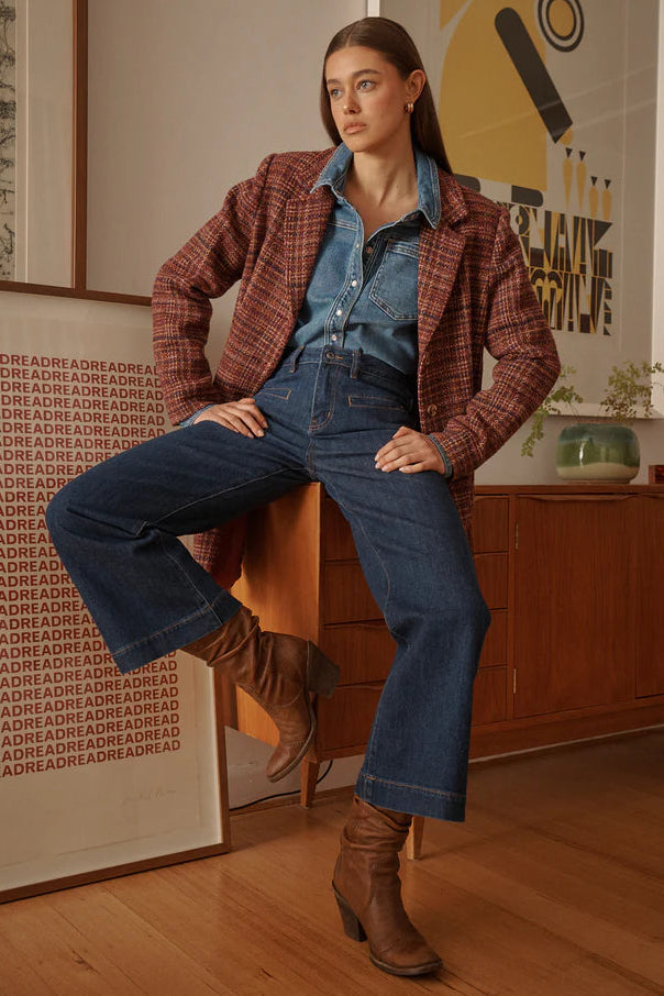 Woman in plaid jacket and blue jeans sitting on a wooden stool in a room with a painting on the wall.