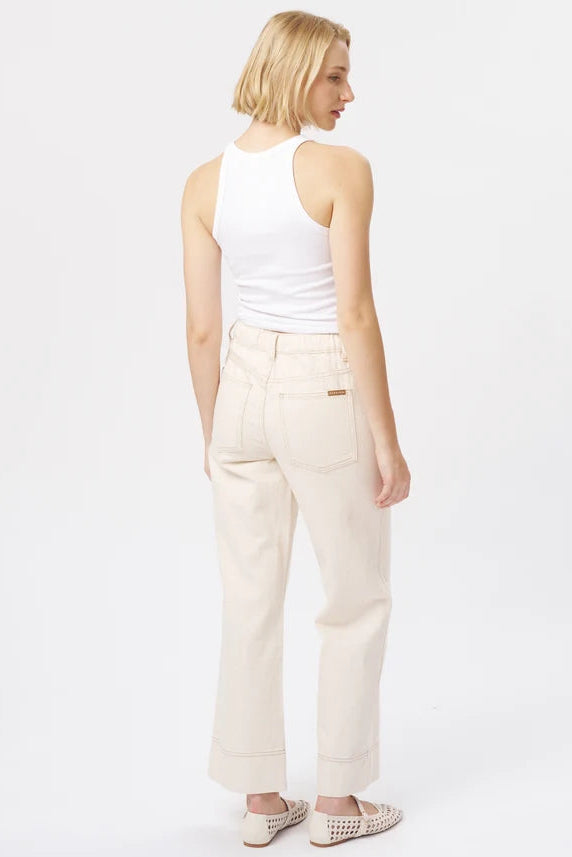 A woman with short blonde hair is shown from the back wearing cream-colored wide-leg jeans, a white tank top, and beige woven slip-on shoes against a plain white studio background.