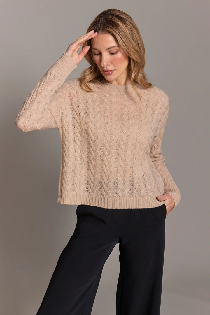 Woman wearing a beige cable knit sweater and black pants against a gray background