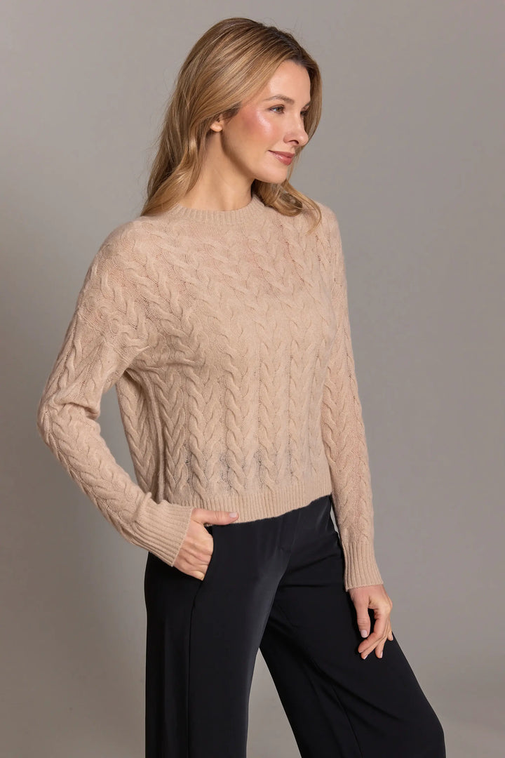 Woman wearing a beige cable knit sweater and black pants against a gray background