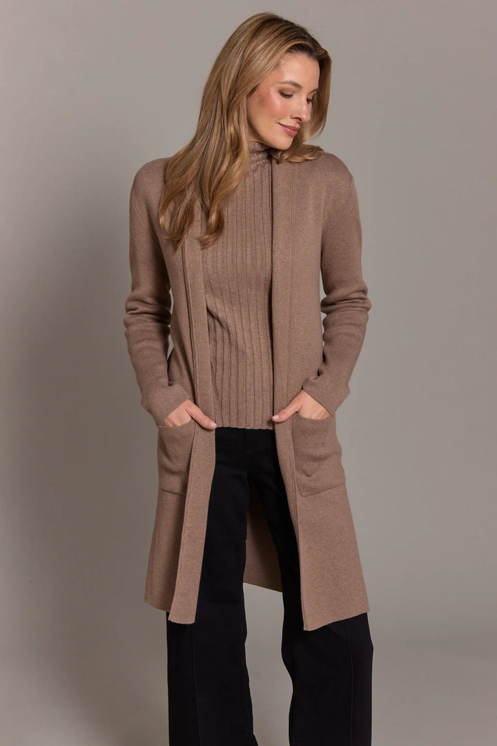 Woman wearing a long brown cardigan over a sweater and black pants on a plain background