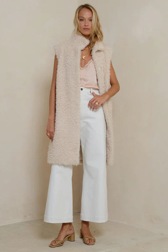 Woman wearing a long beige vest over a pink top and white pants against a plain background