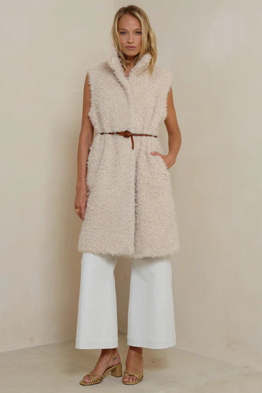 Woman wearing a beige fur vest and white pants against a plain background