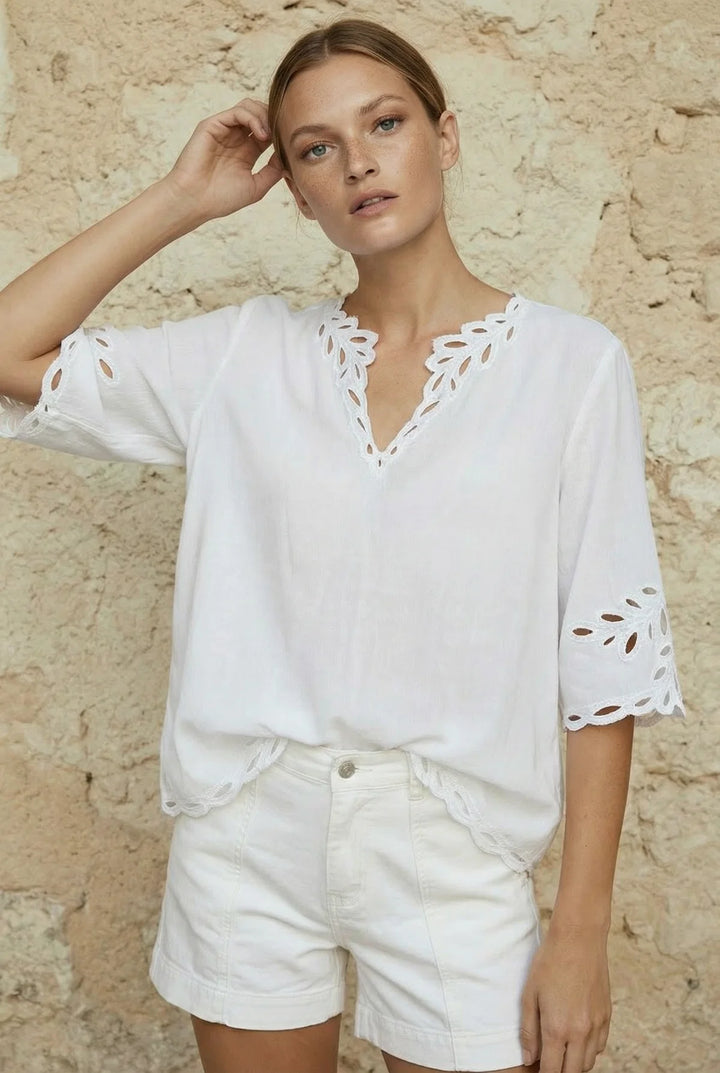A woman wearing a white short-sleeve eyelet blouse with a V-neck and white shorts, standing against a textured beige stone wall.