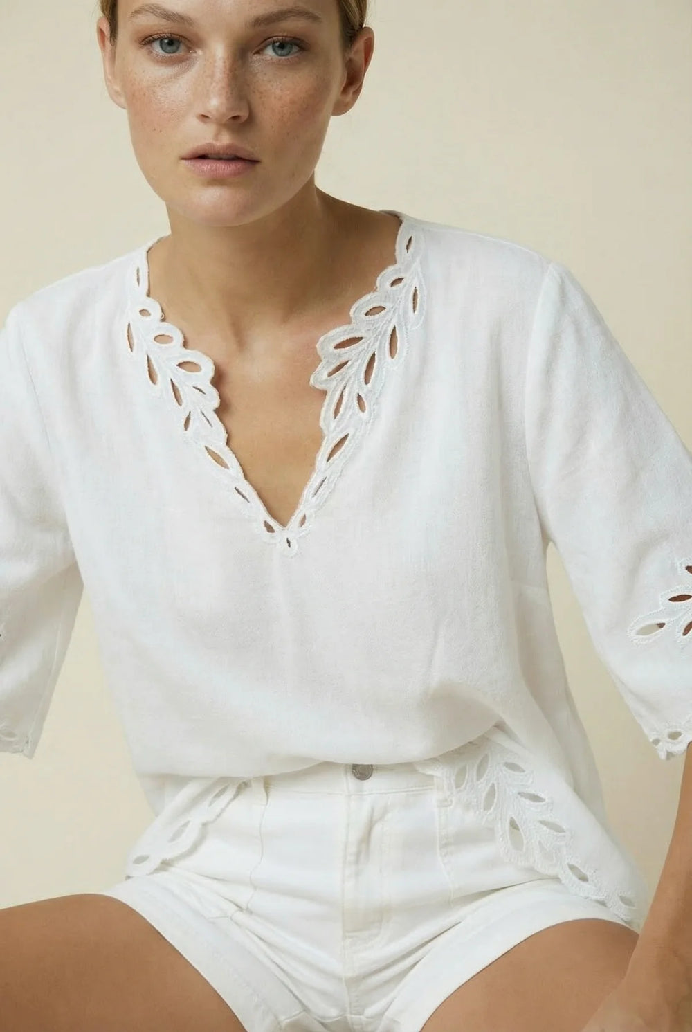 A woman wearing a white short-sleeve top and matching white shorts with eyelet cutout details, photographed against a plain warm beige studio background.