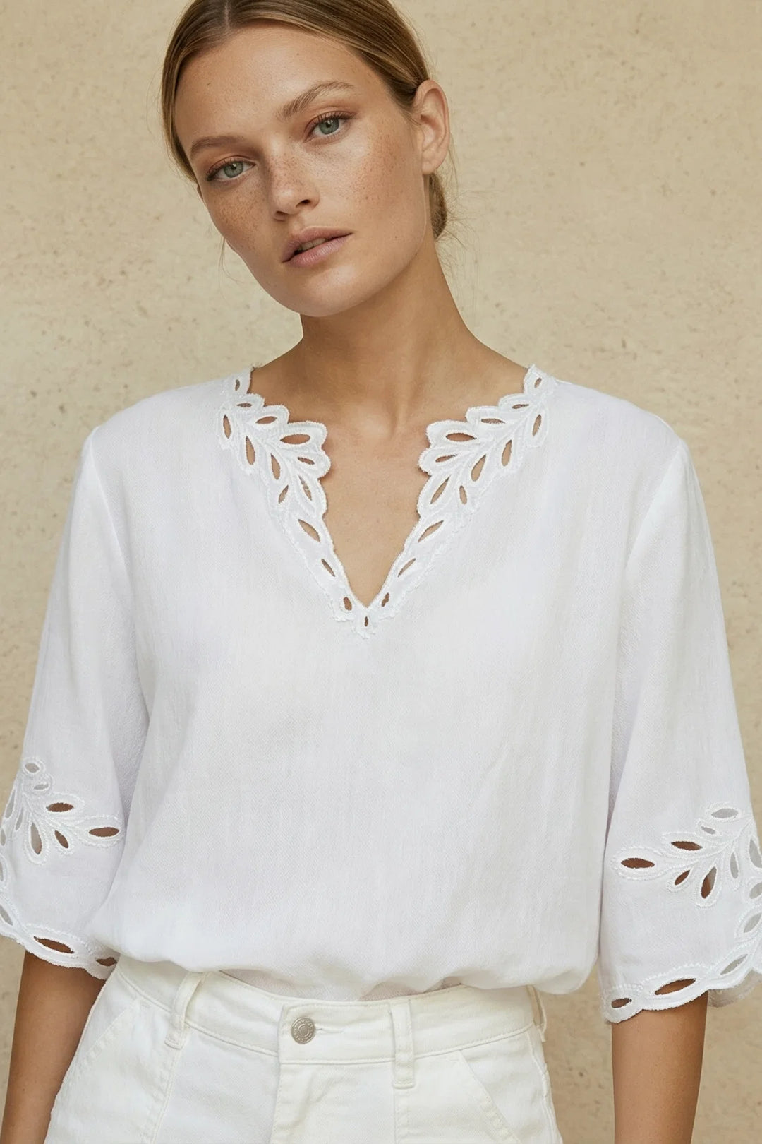 A woman wearing a white eyelet blouse with a V-neck and cutout sleeves, paired with white pants, standing against a textured beige wall.
