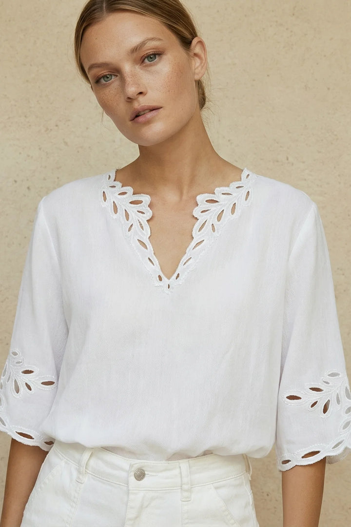 A woman wearing a white eyelet blouse with a V-neck and cutout sleeves, paired with white pants, standing against a textured beige wall.