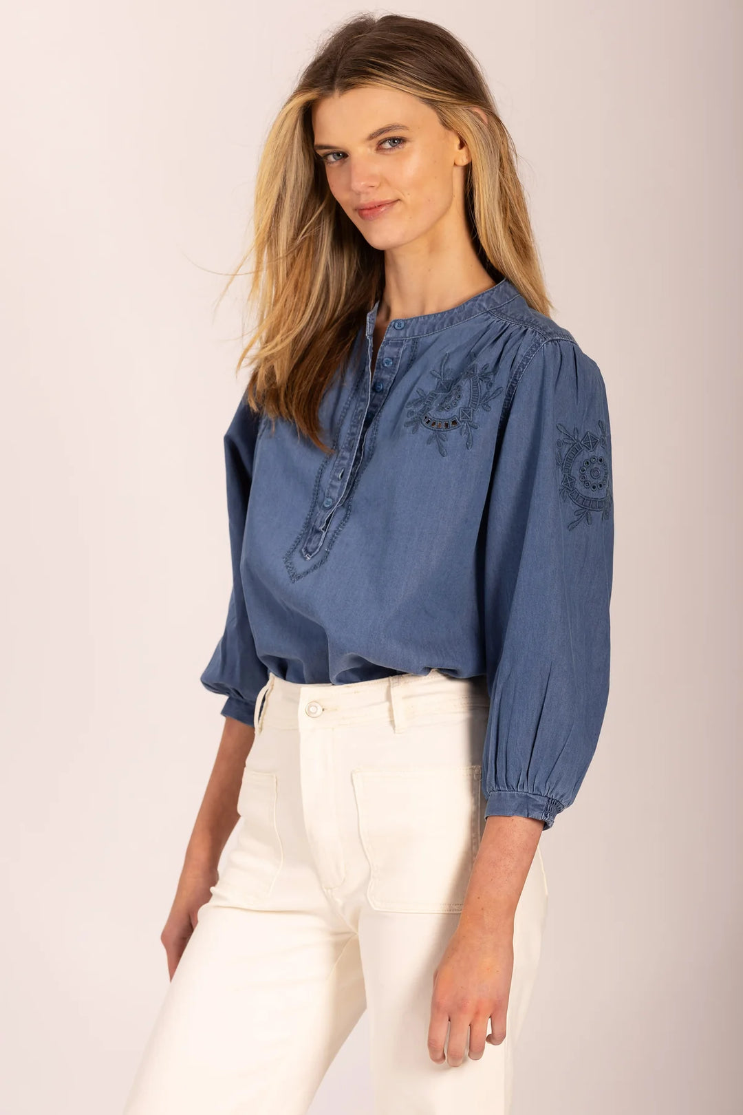 Woman wearing a blue blouse with embroidery and white pants on a plain background