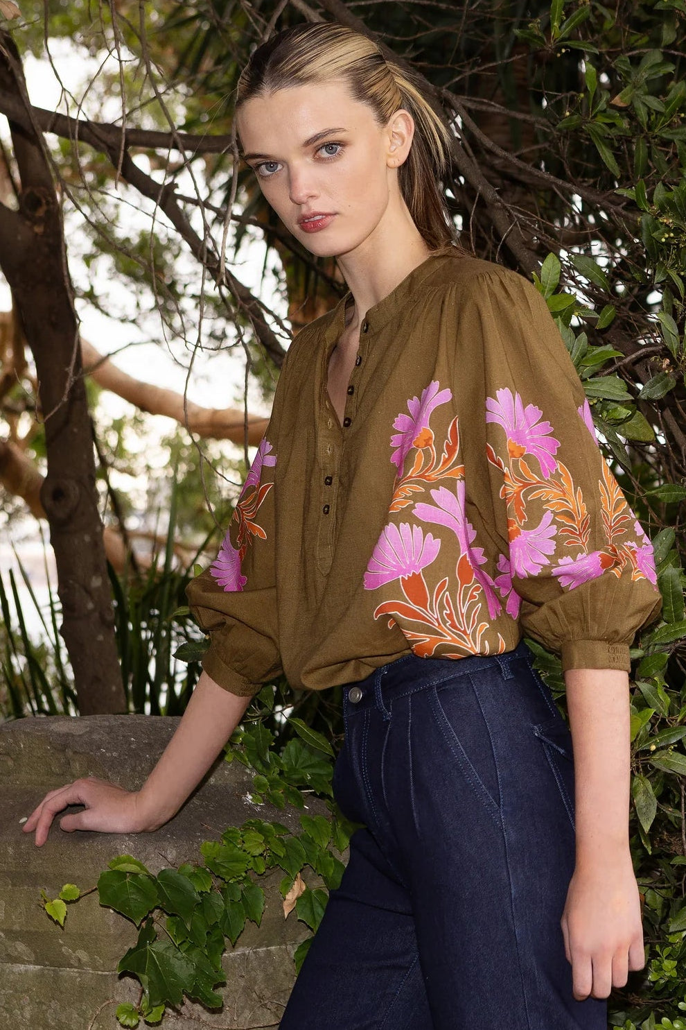 Woman in a floral blouse and dark pants standing in a garden setting