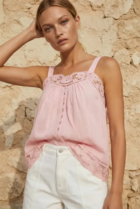 A woman wearing a light pink sleeveless top with delicate lace trim and white pants, posing against a rustic beige stone wall.