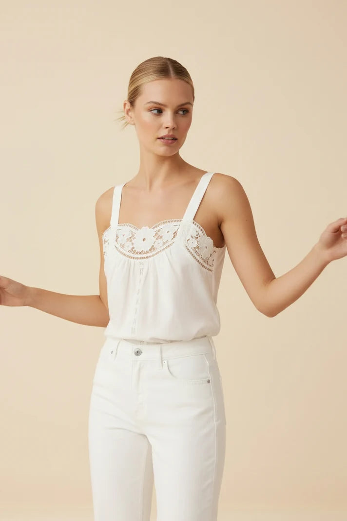 A woman modeling a white sleeveless camisole top with delicate floral lace embroidery across the chest, paired with white pants, against a solid beige studio background.