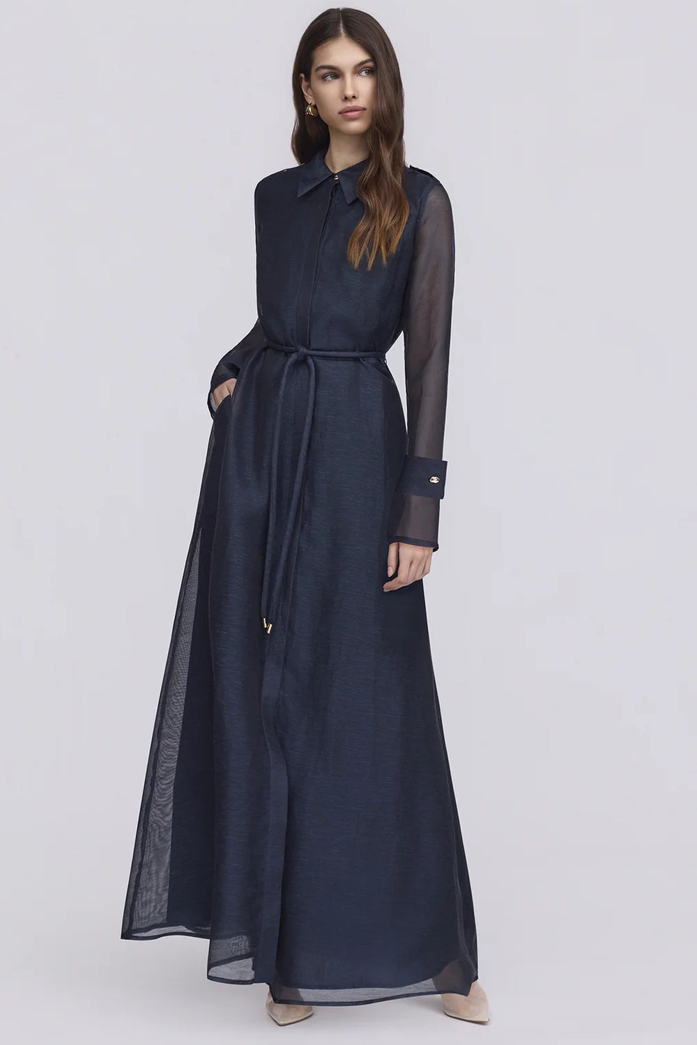 A woman modeling a dark navy blue sleeveless maxi dress with sheer long sleeves, a matching waist tie belt, and side pockets, set against a plain light gray studio background.