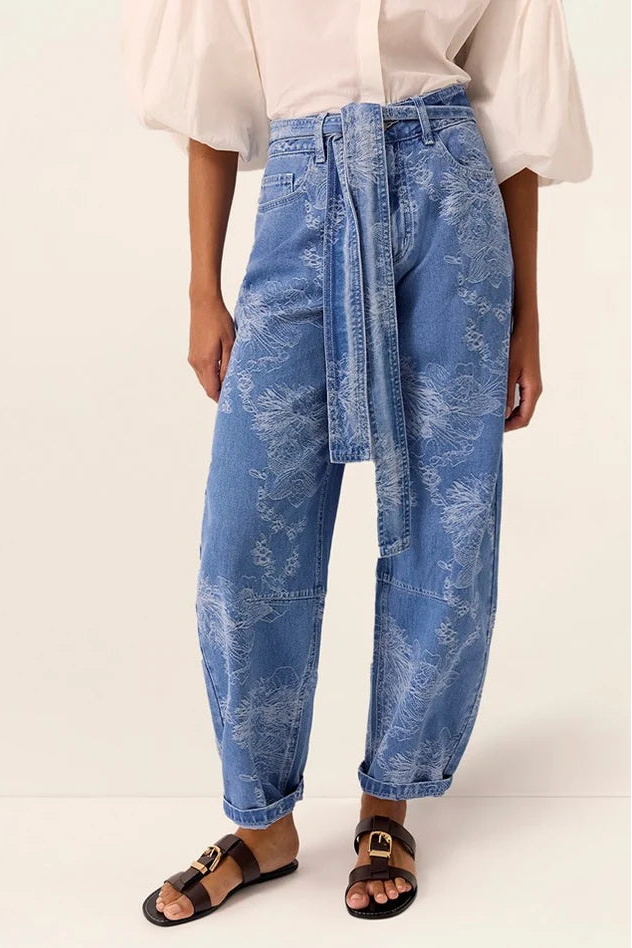 A model wearing high-waisted, wide-leg blue denim jeans with a white abstract floral print and a matching tie belt, styled with a cream blouse and black slide sandals against a light beige studio background.