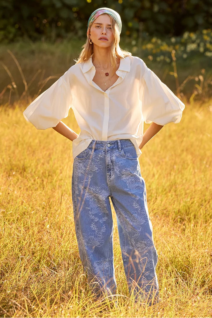 A woman standing in a grassy field at sunset, wearing a loose white button-up blouse, light blue wide-leg jeans with a subtle abstract print, and a multicolor headscarf.