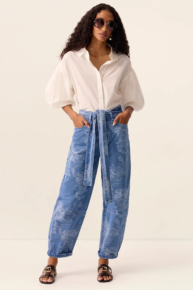 A woman modeling a white long-sleeve blouse and high-waisted, wide-leg blue denim jeans with a tie waist, styled with sunglasses and brown sandals against a plain off-white studio background.