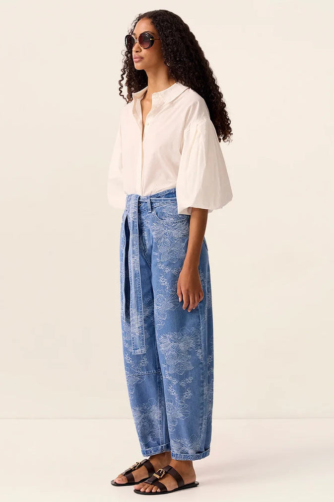 A woman modeling a white short-sleeve blouse and blue patterned wide-leg trousers with black sandals, standing against a plain off-white studio background.