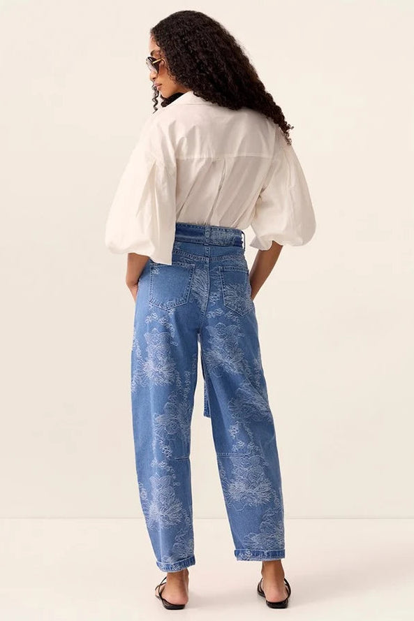 A woman is shown from behind wearing a white puff-sleeve blouse and blue wide-leg jeans with a subtle tonal floral pattern, standing against a plain off-white studio background.