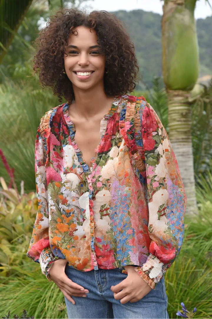 A woman with curly dark hair stands outdoors in a tropical garden setting, wearing a colorful floral print blouse with blue jeans.