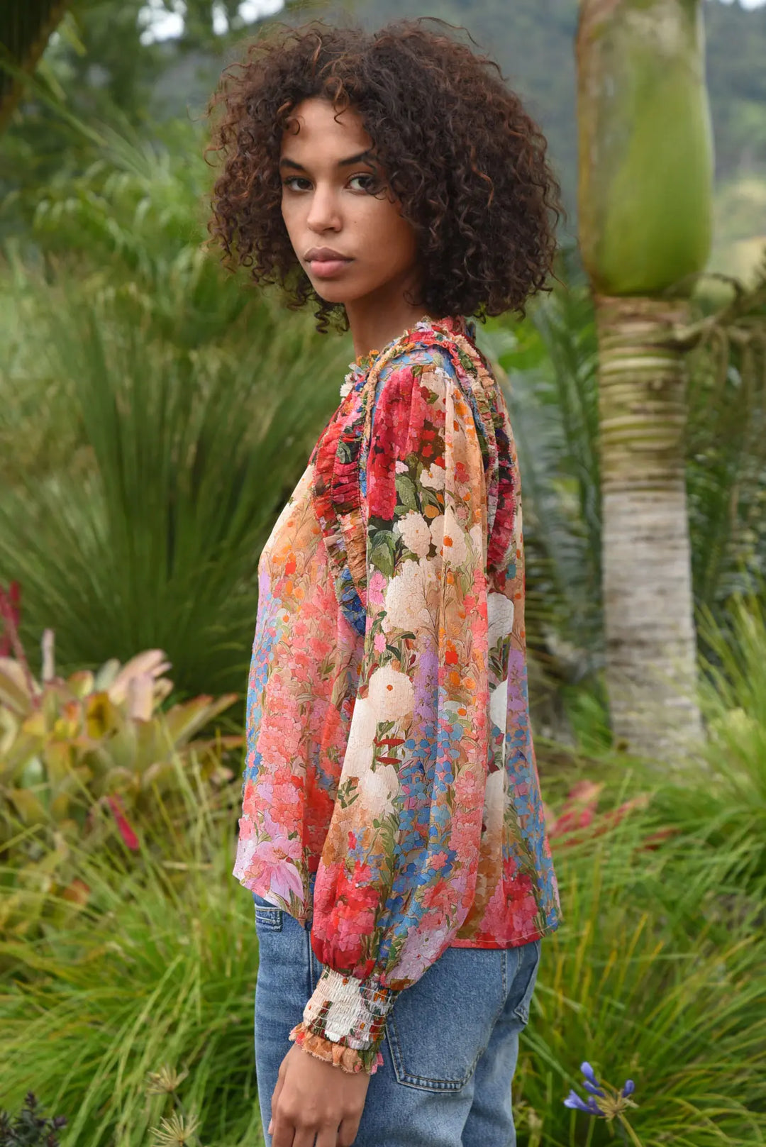 A woman with curly brown hair wearing a colorful floral long-sleeve blouse and blue jeans, standing outdoors in a lush green garden.