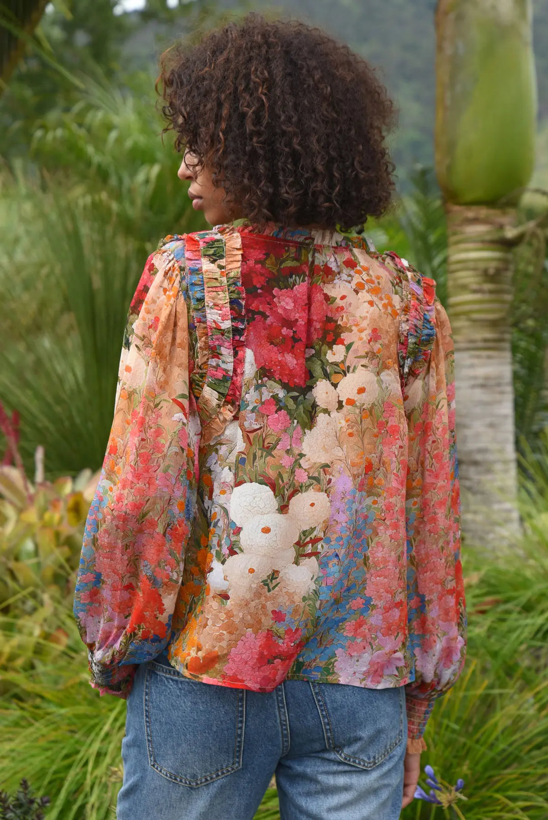 A woman viewed from behind wearing a long-sleeve floral blouse with a multicolor print and blue jeans, standing outdoors in a garden setting.