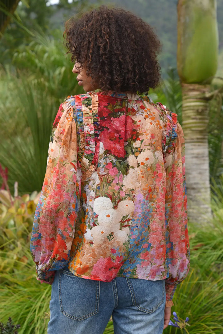 A woman viewed from behind wearing a long-sleeve floral blouse with a multicolor print and blue jeans, standing outdoors in a garden setting.