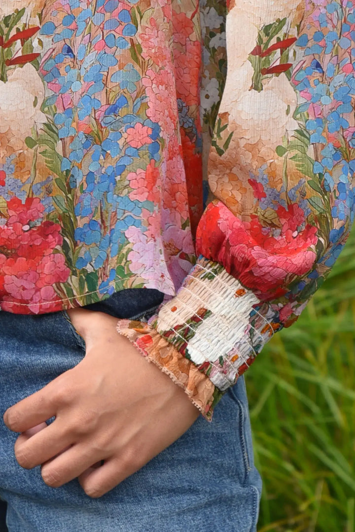 A close-up of a person wearing a long-sleeve blouse with a colorful floral and abstract print, featuring a smocked cuffed sleeve and blue jeans, with green grass in the background.