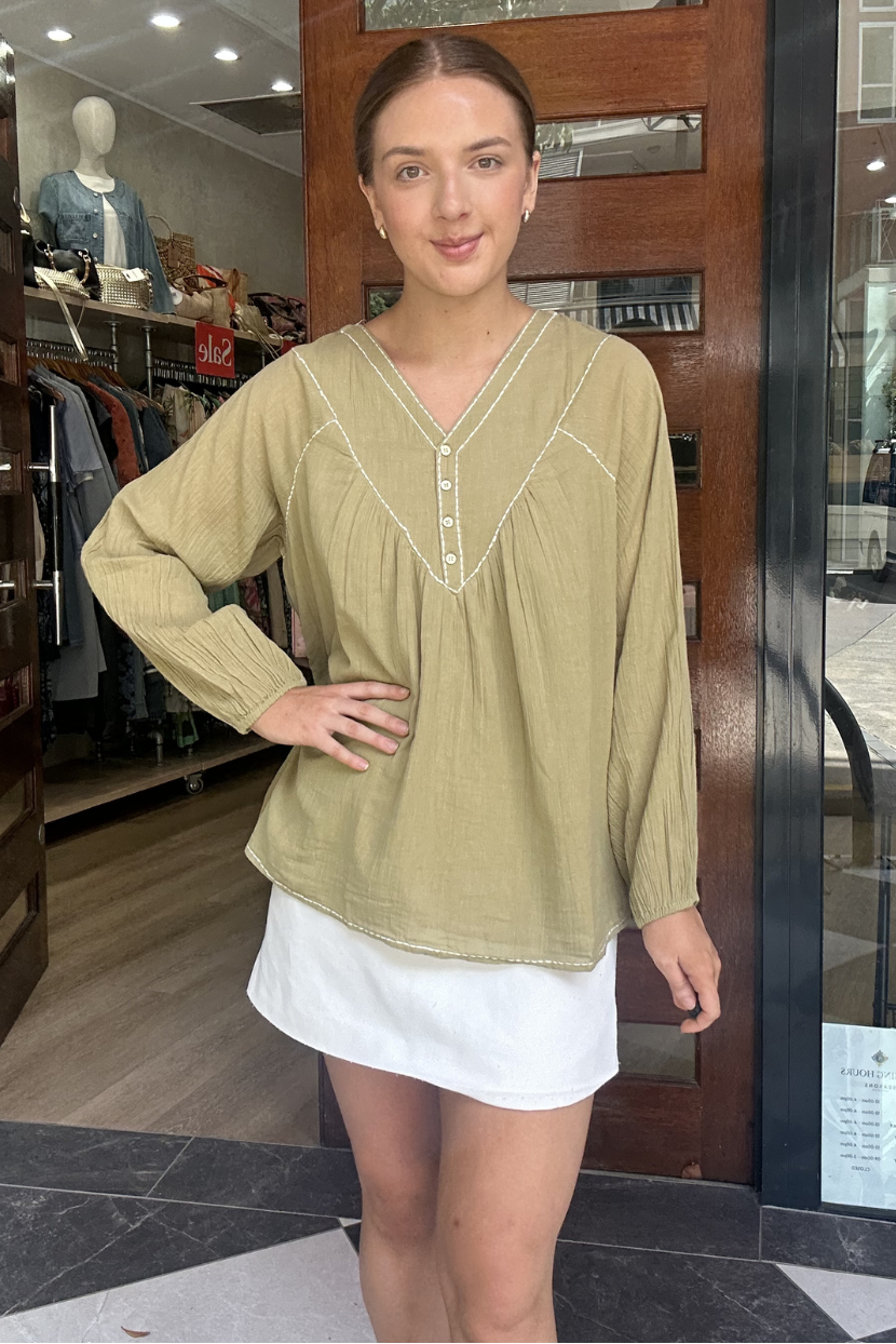Woman standing in front of a store entrance wearing a green top and white skirt.