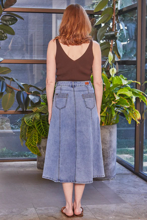 Woman wearing a brown top and blue denim skirt standing in front of a glass door with plants.