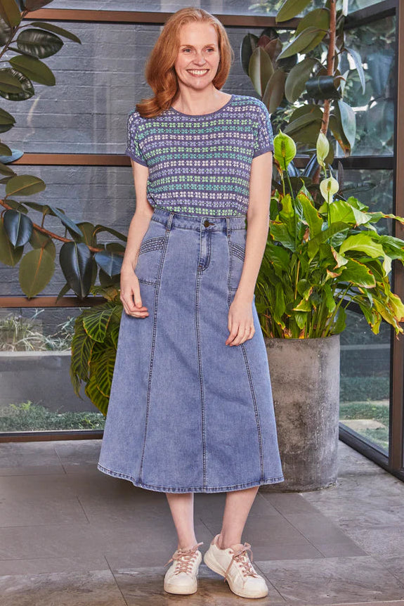 Woman in a patterned top and denim skirt standing indoors with plants in the background