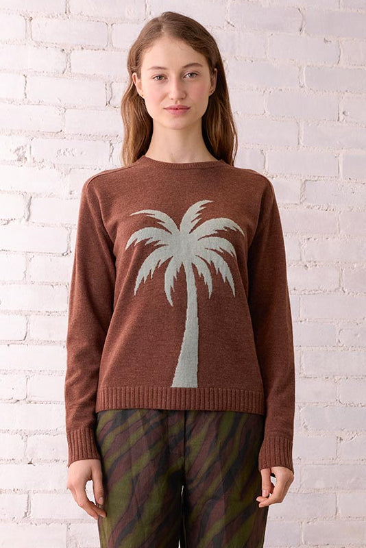 Woman wearing a brown sweater with a palm tree design in a room with a white brick wall and wicker chair.