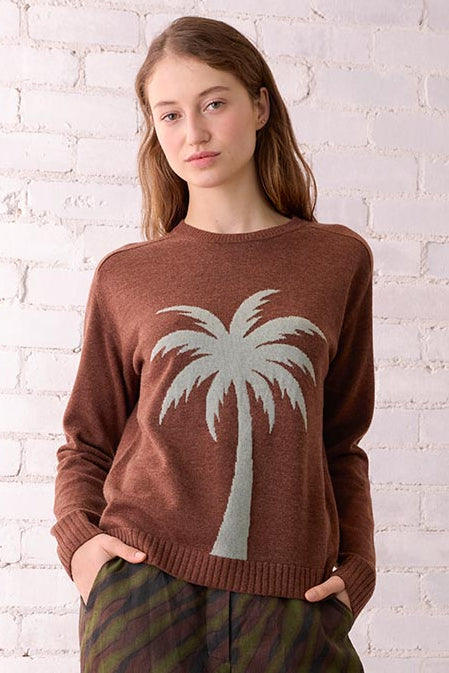 Woman wearing a brown sweater with a palm tree design and striped pants, standing in a room with a white brick wall and wicker chair.