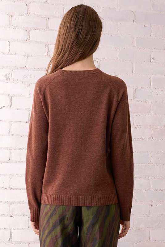 Person wearing a brown sweater and patterned pants standing in a room with a white brick wall and wicker chair.