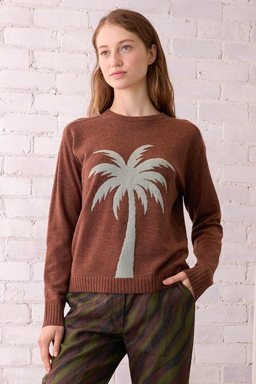 Woman wearing a brown sweater with a palm tree design and striped pants, standing in a room with a white brick wall and wicker chair.