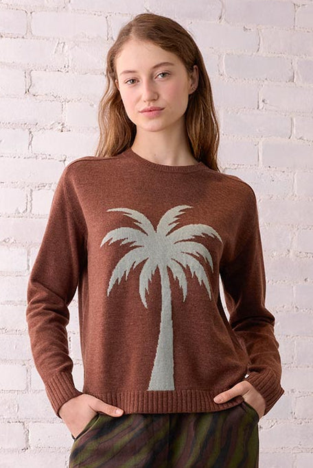 Woman wearing a brown sweater with a palm tree design, standing in a room with a white brick wall and wicker chair.
