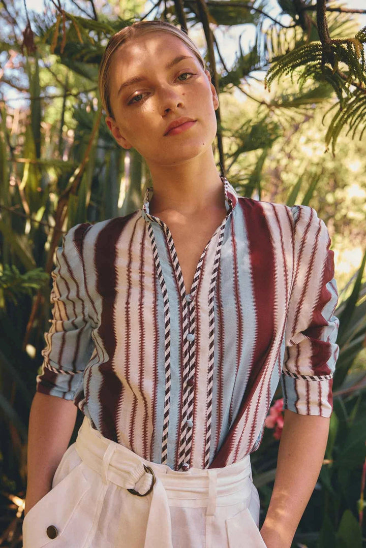 Woman wearing a striped shirt and white pants standing in front of green foliage