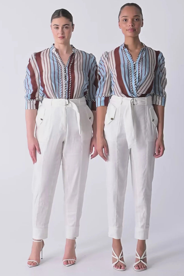 Two models standing side by side wearing matching outfits consisting of white high-waisted cropped trousers with tie details and striped button-up shirts in blue, brown, and white.