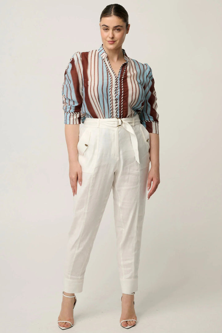 A woman modeling a long-sleeve button-up shirt with vertical stripes in blue, red, and white, paired with high-waisted white trousers and beige heeled sandals, against a plain light gray studio background.