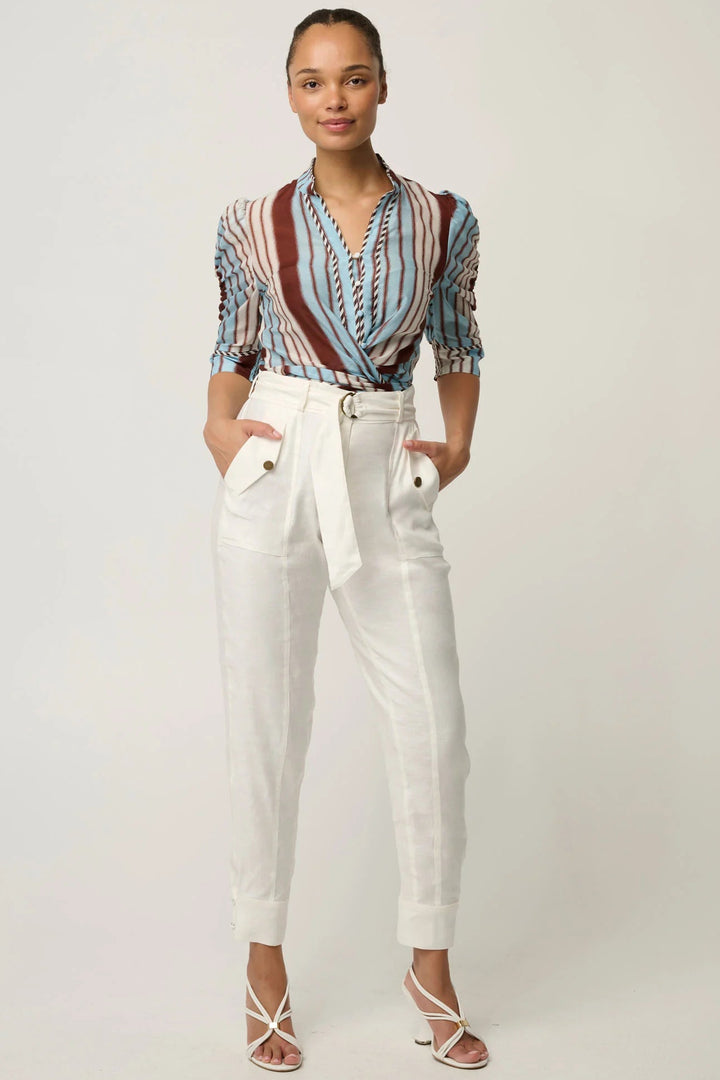 A woman modeling a short-sleeve button-up blouse with vertical multicolor stripes in blue, red, and brown, paired with white high-waisted trousers and strappy heels, against a light gray studio background.
