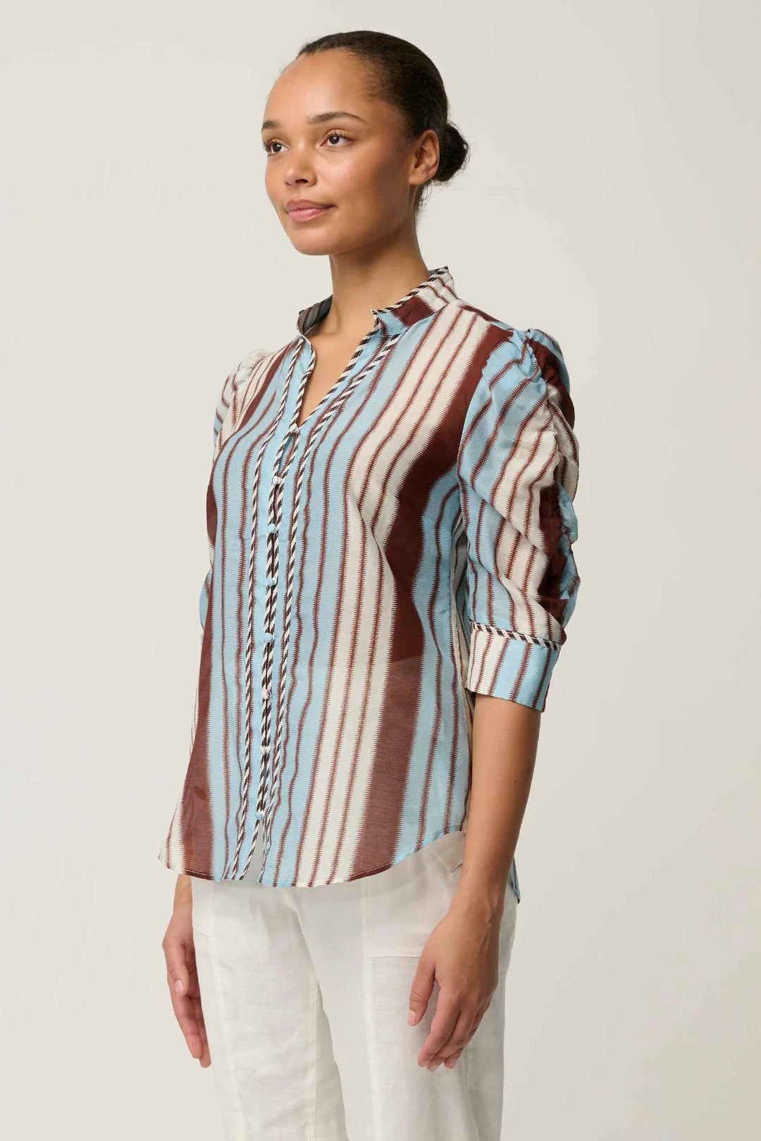 A woman wearing a long-sleeve button-up blouse with vertical stripes in light blue, brown, and white, paired with white pants, photographed against a plain light gray studio background.