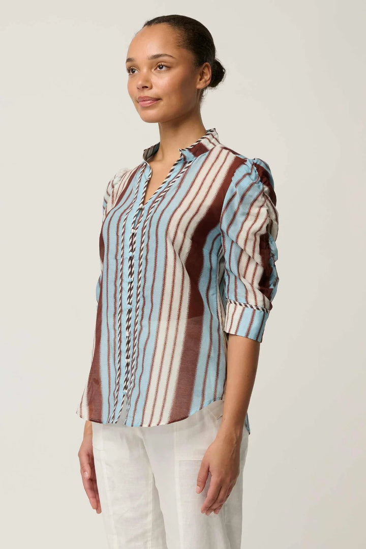 A woman wearing a long-sleeve button-up blouse with vertical stripes in light blue, brown, and white, paired with white pants, photographed against a plain light gray studio background.