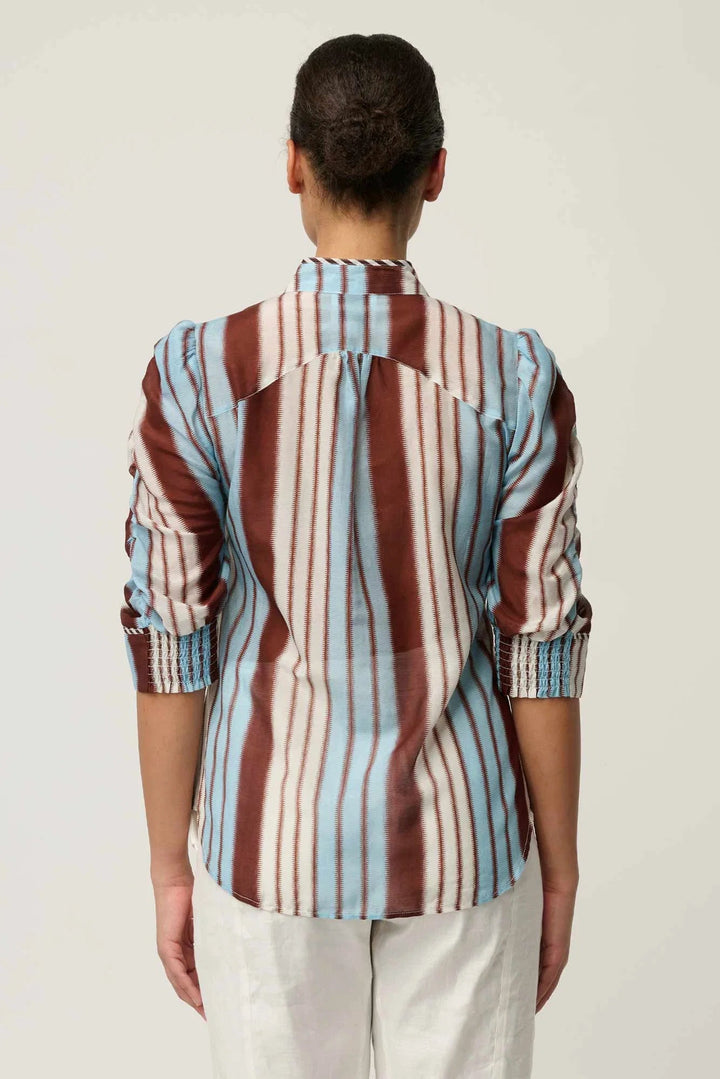 A woman viewed from the back wearing a long-sleeve button-up shirt with vertical stripes in brown, light blue, and off-white, paired with off-white trousers, against a plain light gray background.