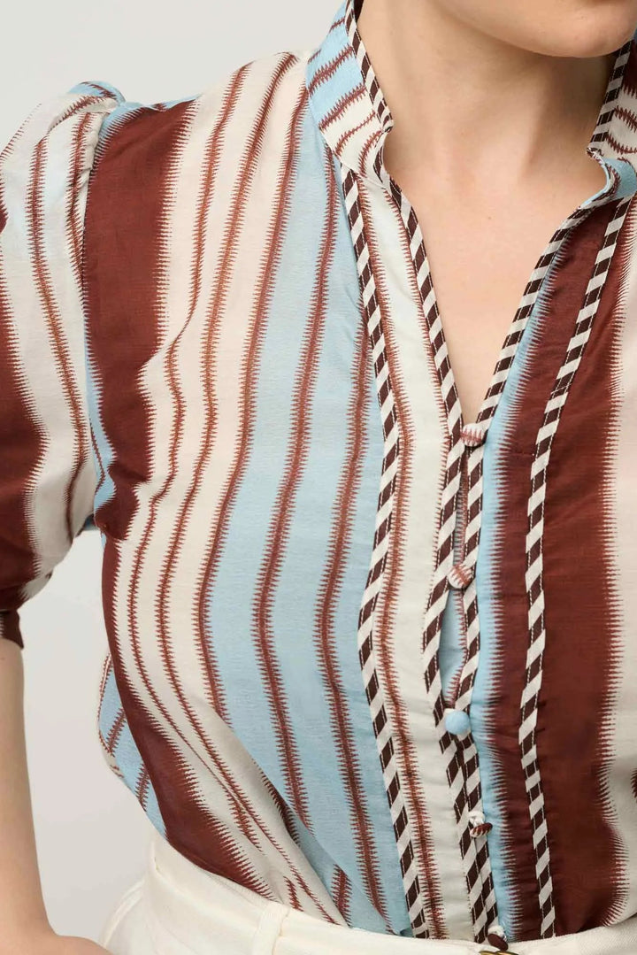 A close-up of a woman wearing a short-sleeve button-front blouse with vertical wavy stripes in brown, cream, and light blue, plus a striped tie-style placket.