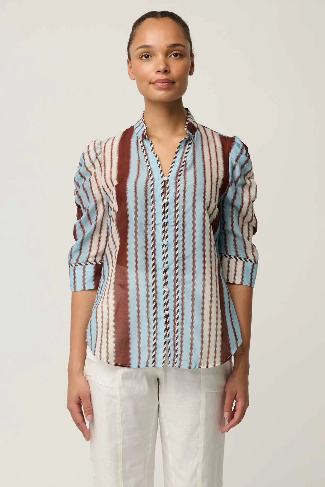 A woman modeling a long-sleeve button-up shirt with vertical stripes in brown, light blue, beige, and white, paired with white pants, against a plain light gray background.