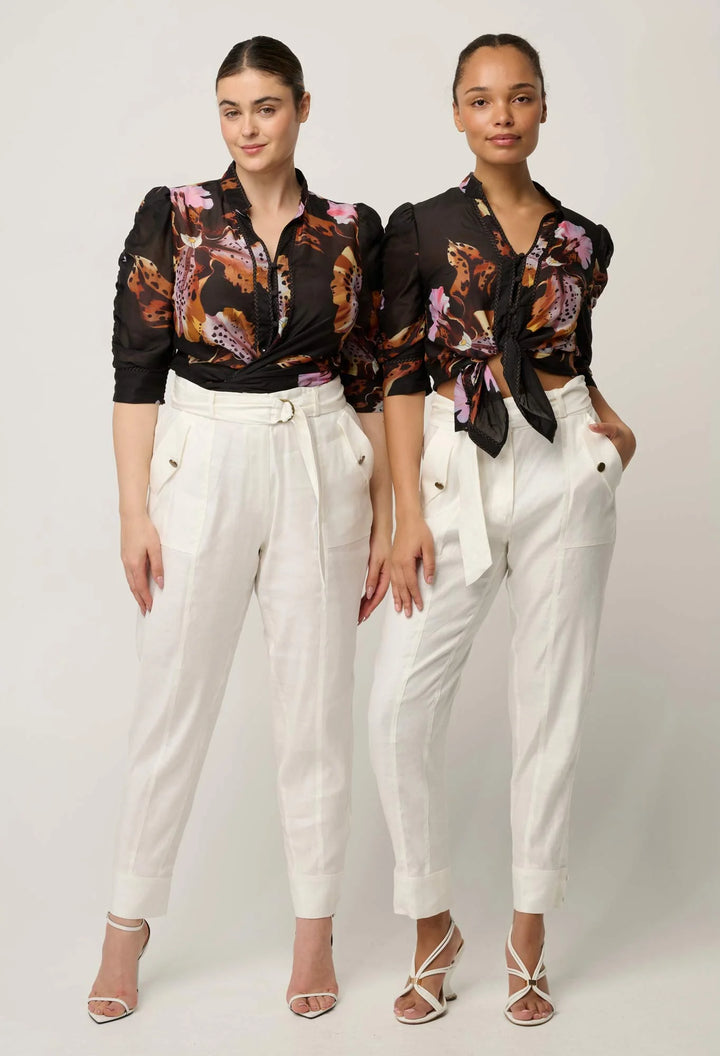 Two models wearing matching outfits consisting of black long-sleeve button-up shirts with a colorful floral print and white high-waisted trousers, standing against a light gray studio background.