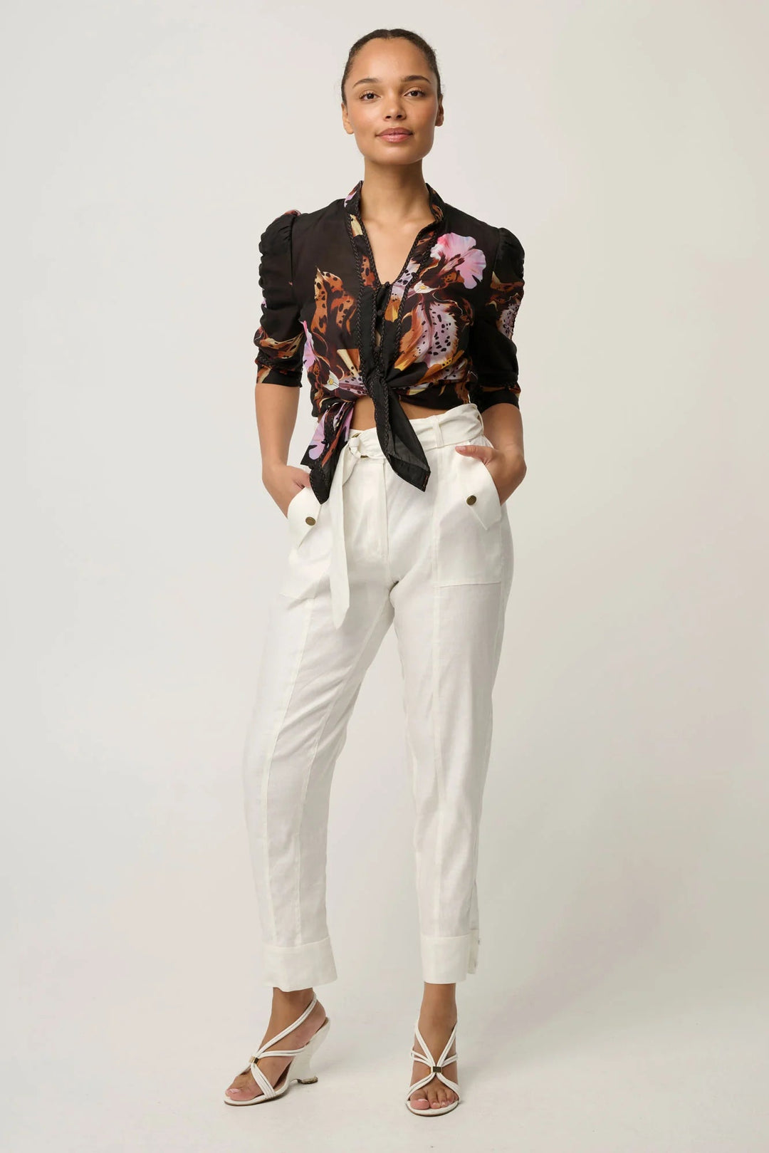 A model stands against a light gray studio background wearing a black floral long-sleeve blouse tied at the waist and white high-waisted cropped trousers with rolled cuffs.