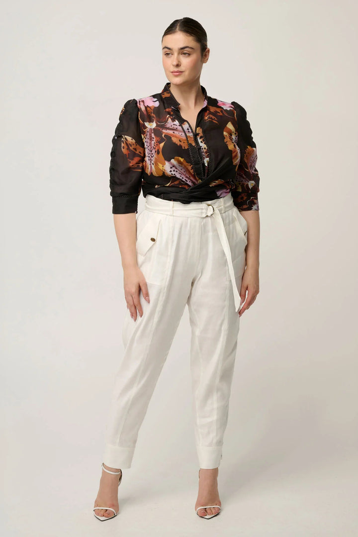 A woman modeling a black long-sleeve button-up shirt with a colorful abstract floral print, paired with white high-waisted trousers and nude high-heeled sandals, against a light gray studio background.
