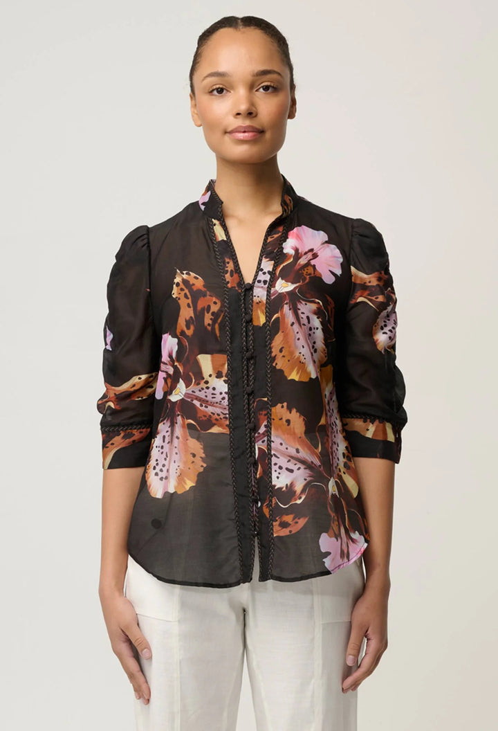 A woman modeling a black sheer button-up blouse with a brown, orange, and pink butterfly print, paired with white trousers, against a light gray studio background.