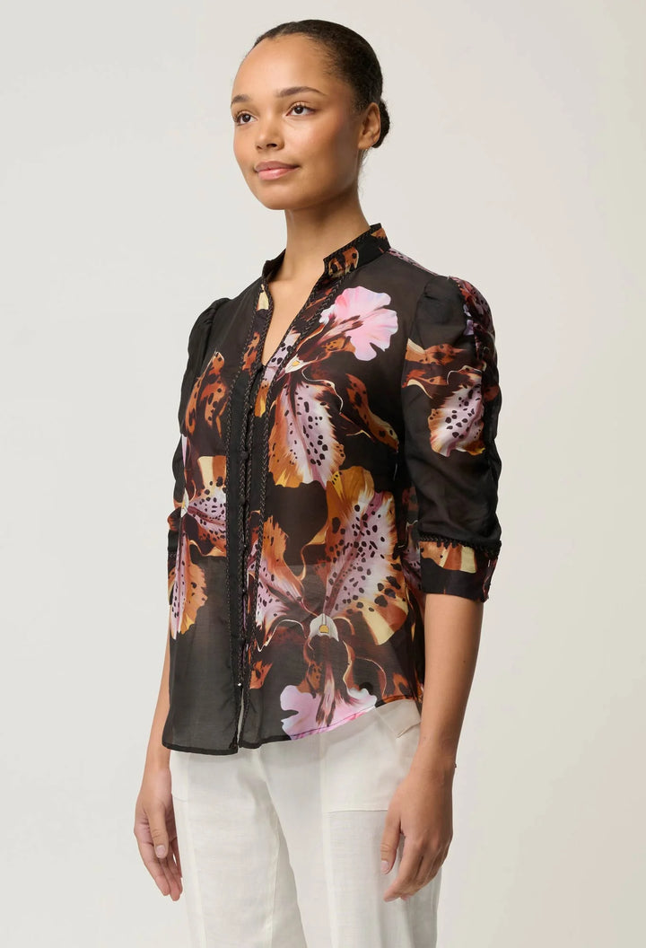 A woman modeling a sheer black button-up blouse with a vibrant floral print in pink, orange, and brown, paired with white trousers, against a plain light gray studio background.
