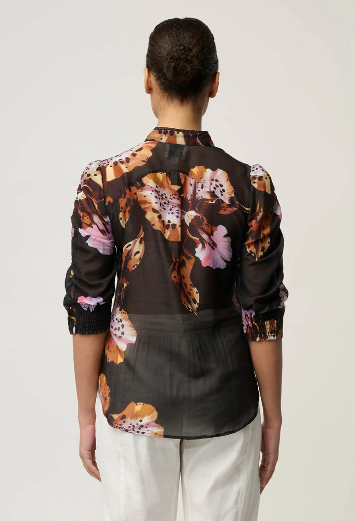 A woman viewed from the back wearing a sheer black blouse with a large floral print in brown, orange, and pink, paired with white pants, against a light gray studio background.