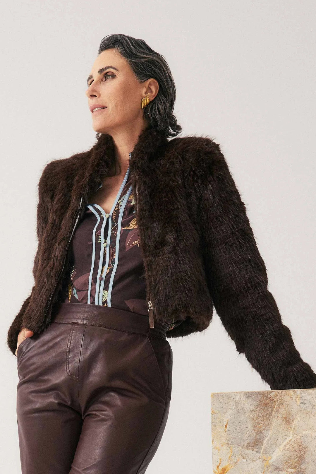 Woman wearing a brown fur jacket and leather pants against a plain background